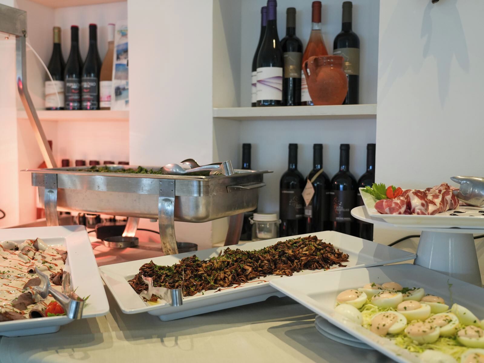 A buffet with various dishes, including meat, vegetables, and eggs, and a selection of wine bottles on shelves in the background.