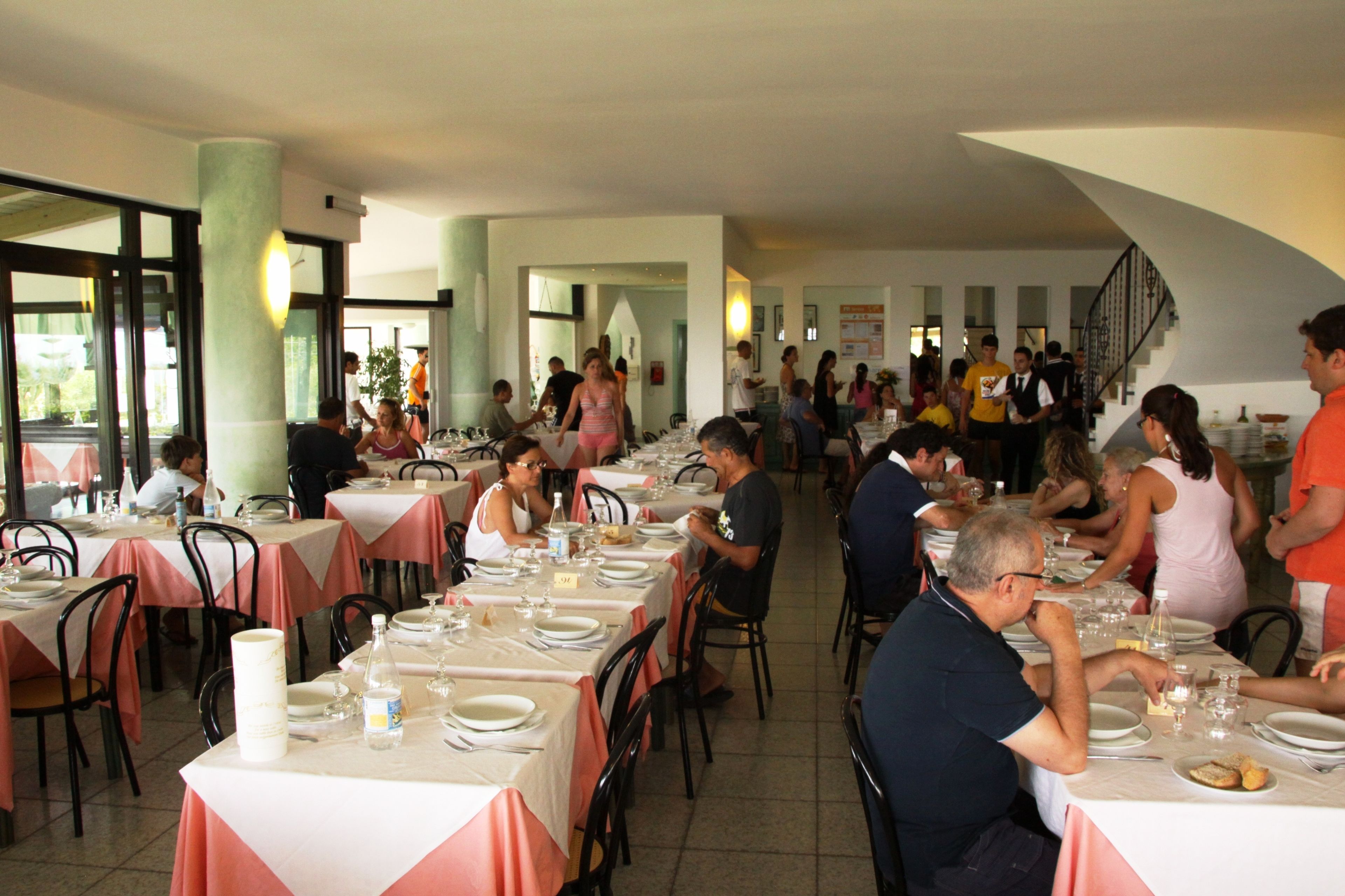 Un vaste espace dédié à la salle à manger, avec de nombreuses tables et chaises, de grandes fenêtres et un escalier menant à un niveau supérieur.