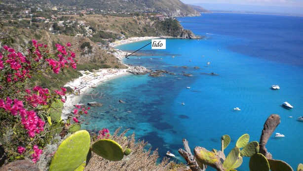 A coastal landscape with a beach, clear blue sea, boats, and a mountainous backdrop.