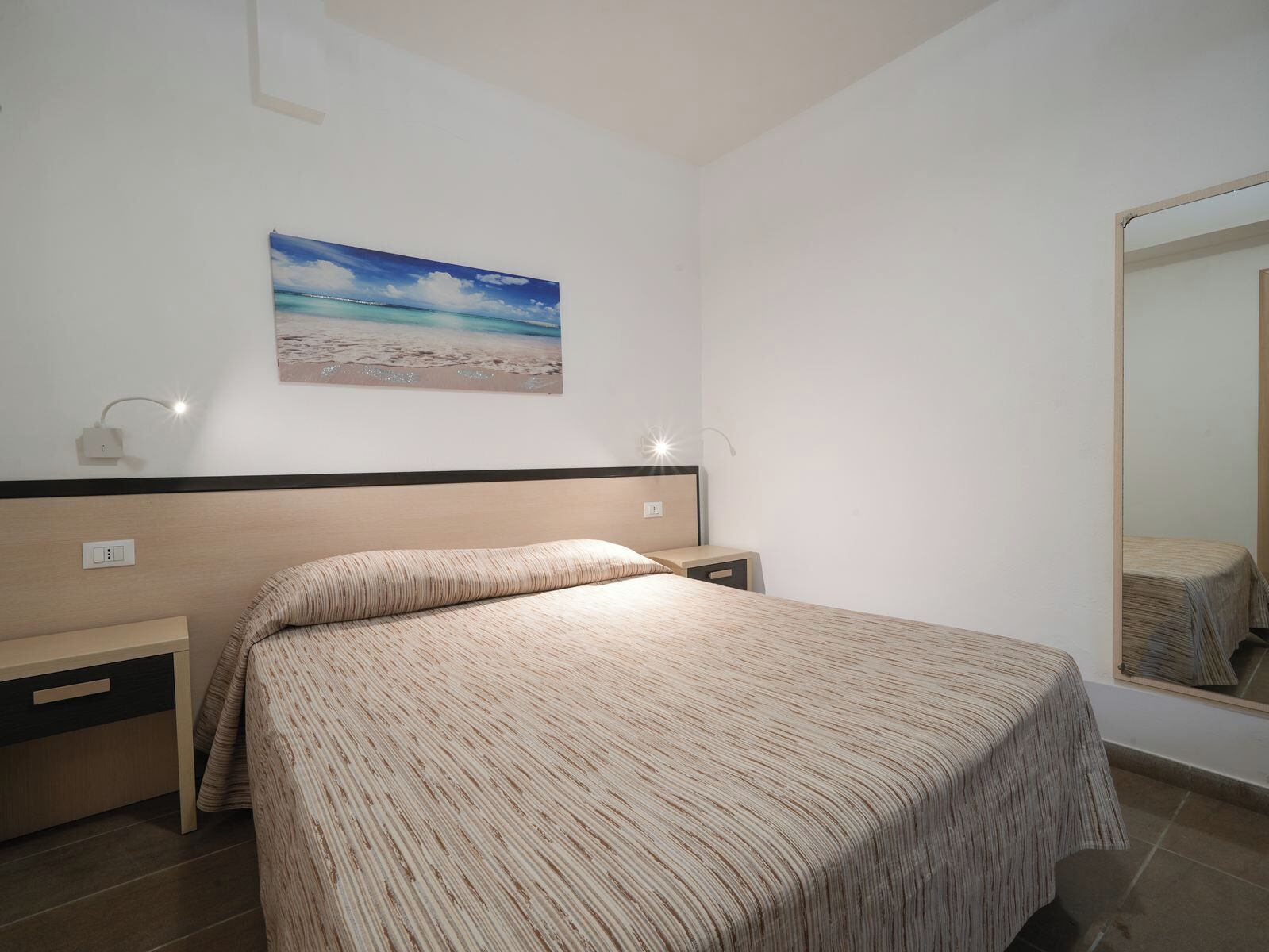 Een slaapkamer met een bed, nachtkastje, een wandlamp, een schilderij van een strand en een spiegel.