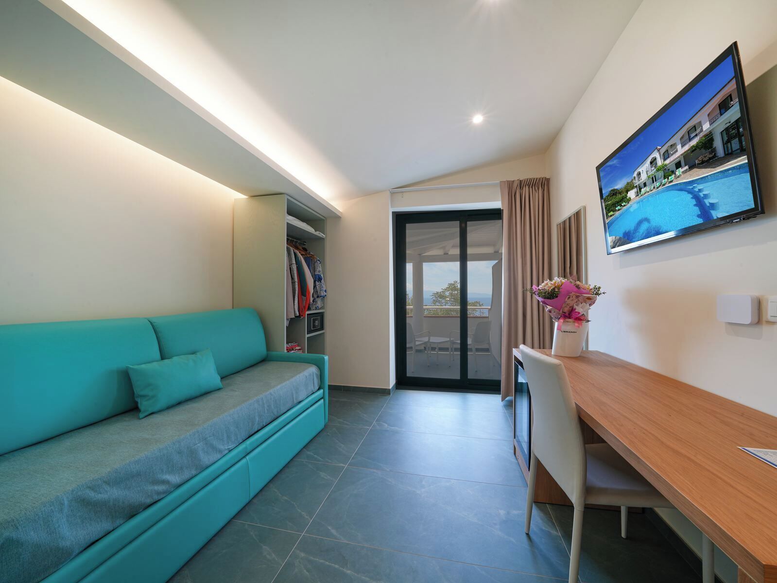 Una habitación de hotel moderna con un sofá color turquesa, un escritorio de madera, un televisor en la pared y un balcón con vista a la piscina.