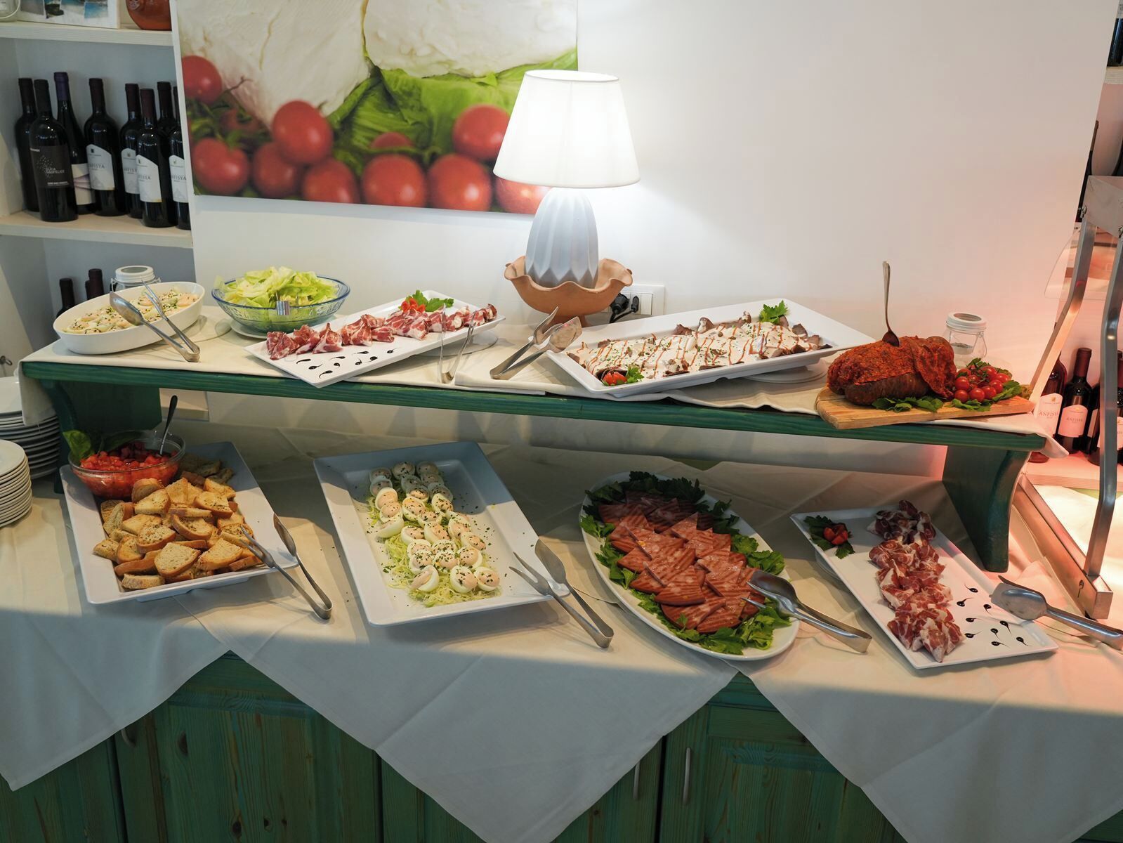 A buffet table with various dishes, including meats, cheeses, and salads, under a table lamp.