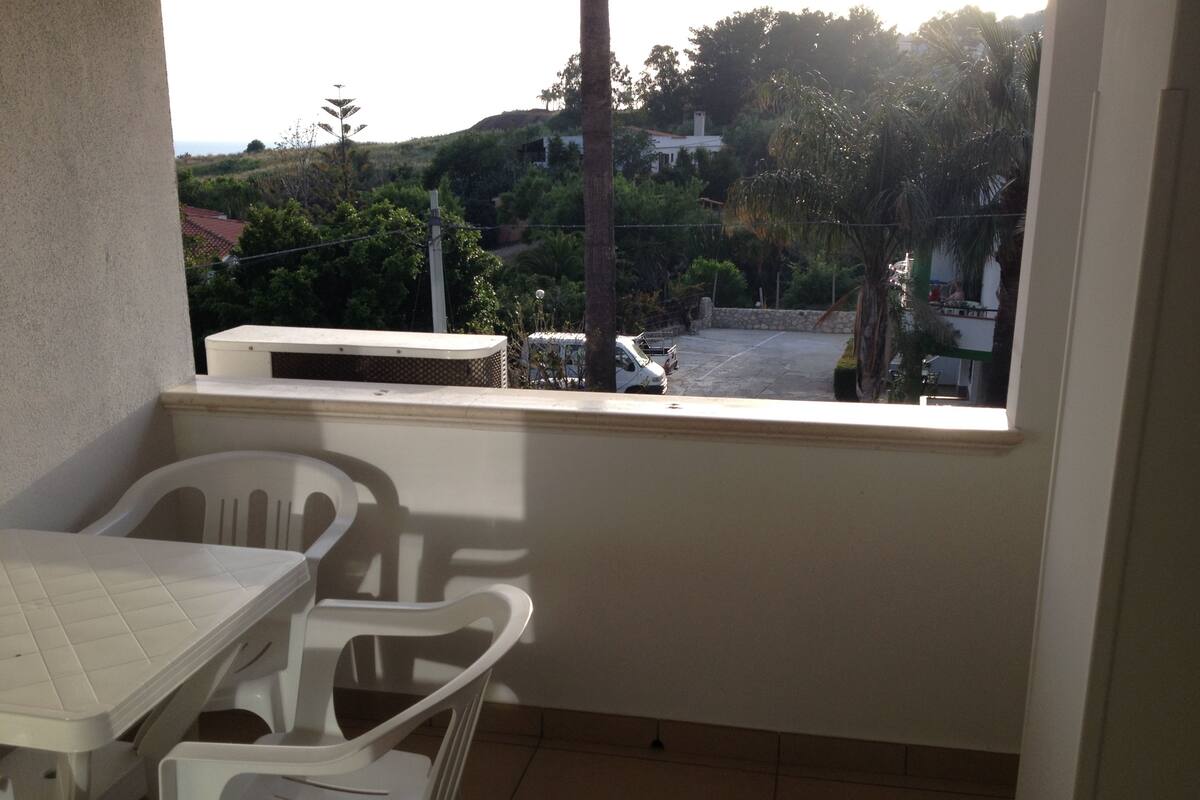 A balcony with a white table and chairs, a view of a parking area, and a building in the background.