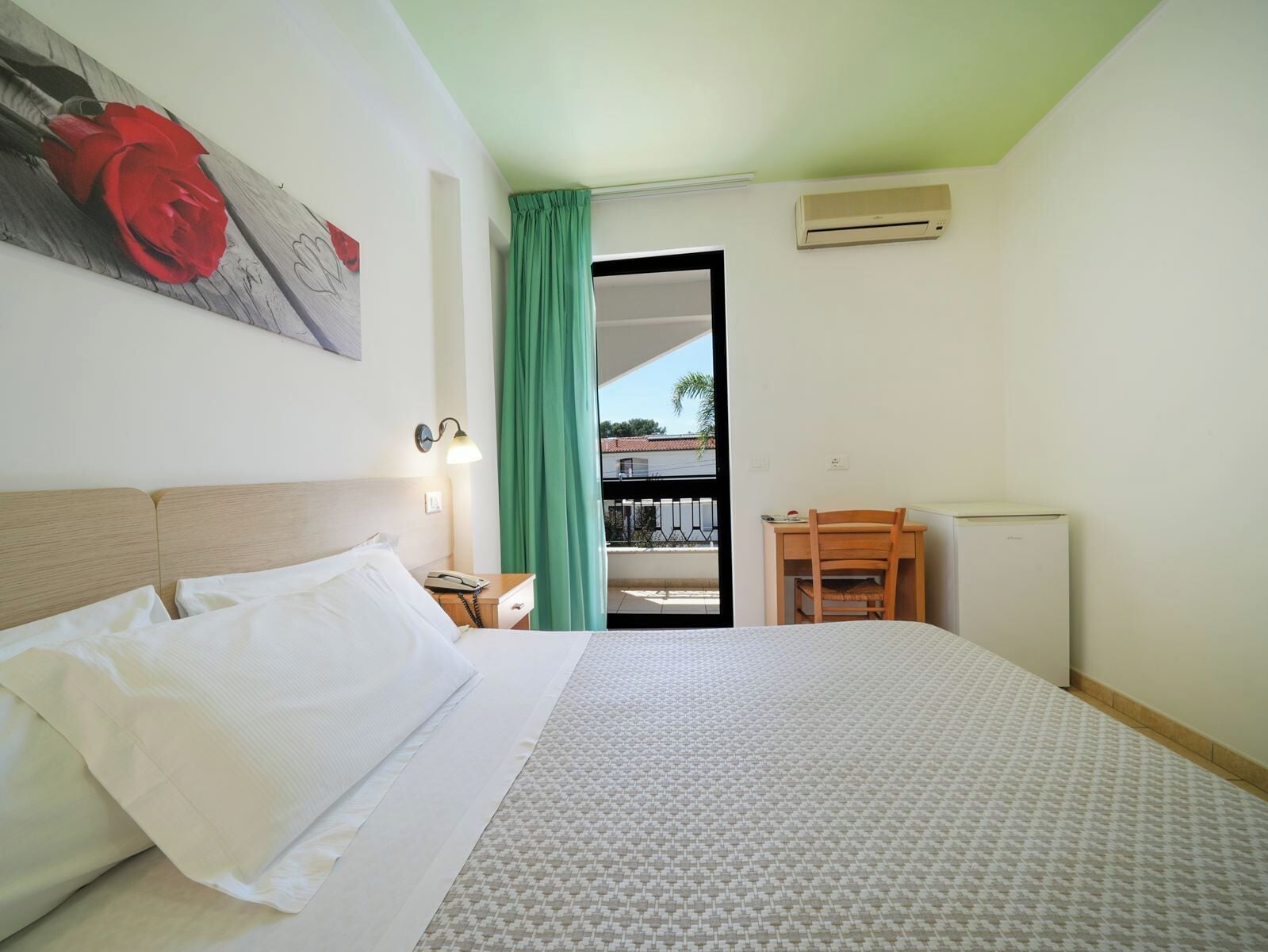 Habitación de hotel con cama, escritorio, silla, mesita de noche, lámpara, un cuadro y vistas al exterior.