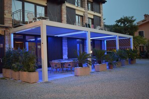 Uma área de pátio externa com espaço de estar coberto, plantas em vasos e um edifício ao fundo.