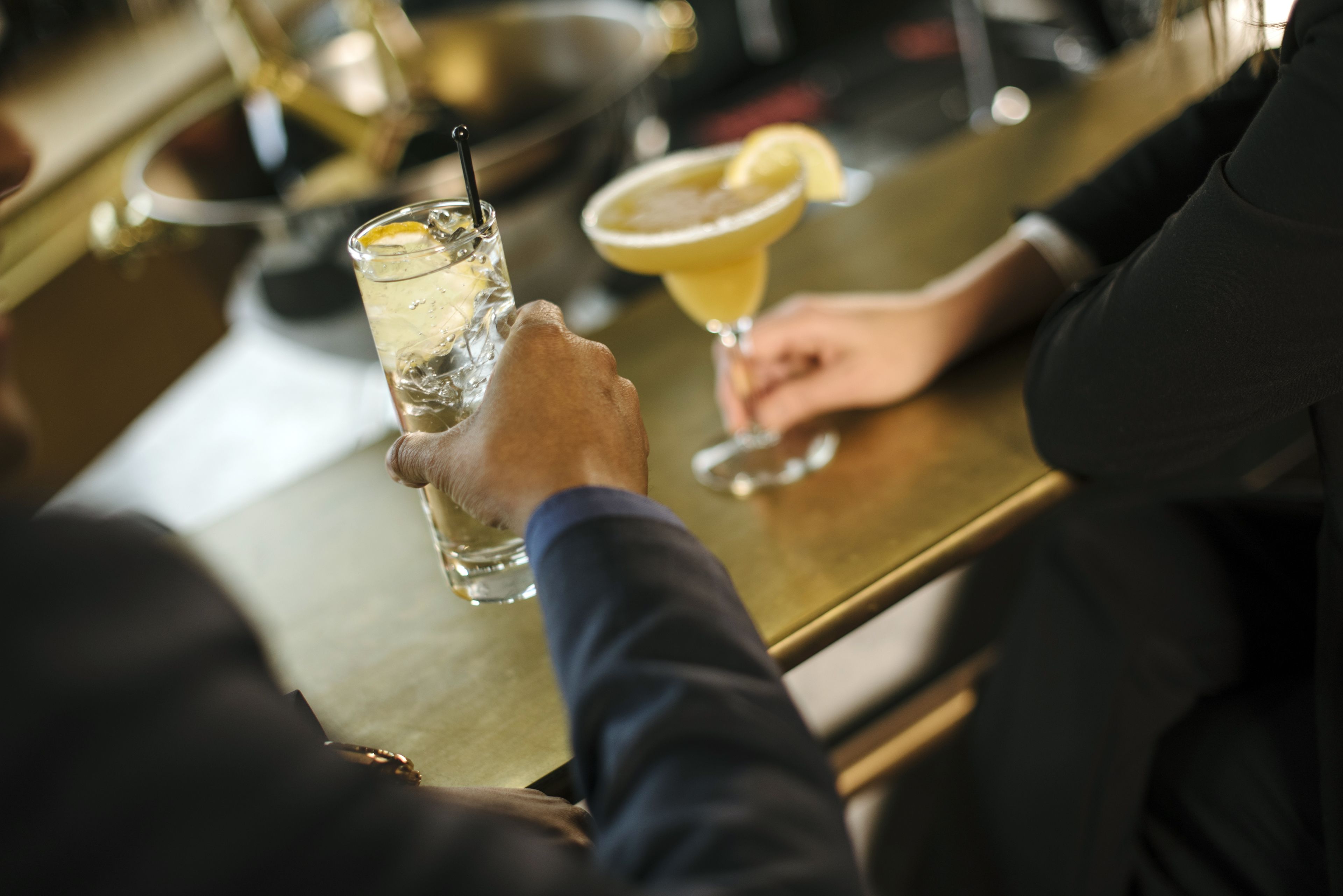 Un bar con una persona sosteniendo un vaso de una bebida transparente con hielo y una rodaja de limón, y otro vaso con una bebida amarilla adornada con una rodaja de limón.