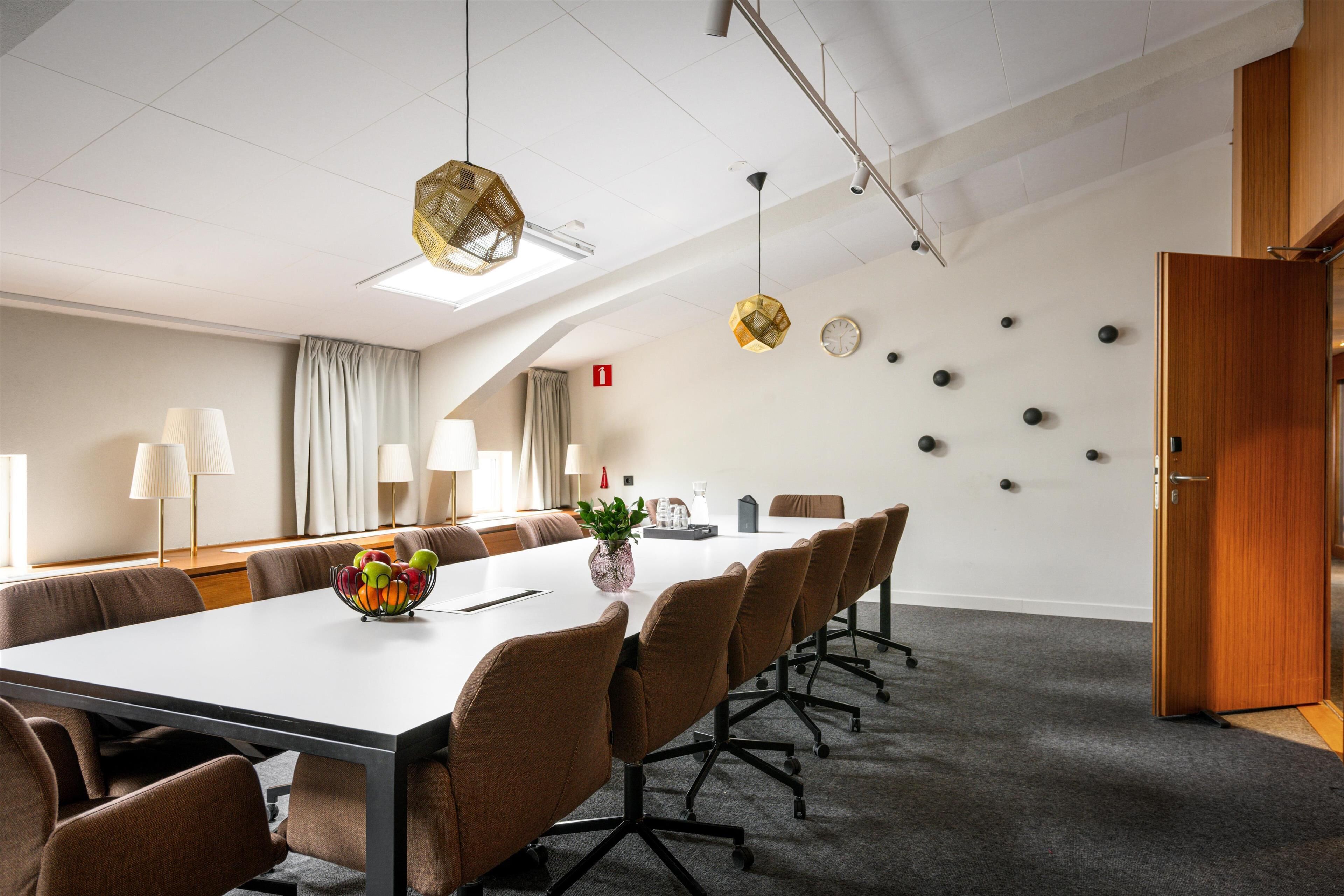 Un salón de conferencias con una larga mesa, sillas marrones y un centro de mesa con planta.