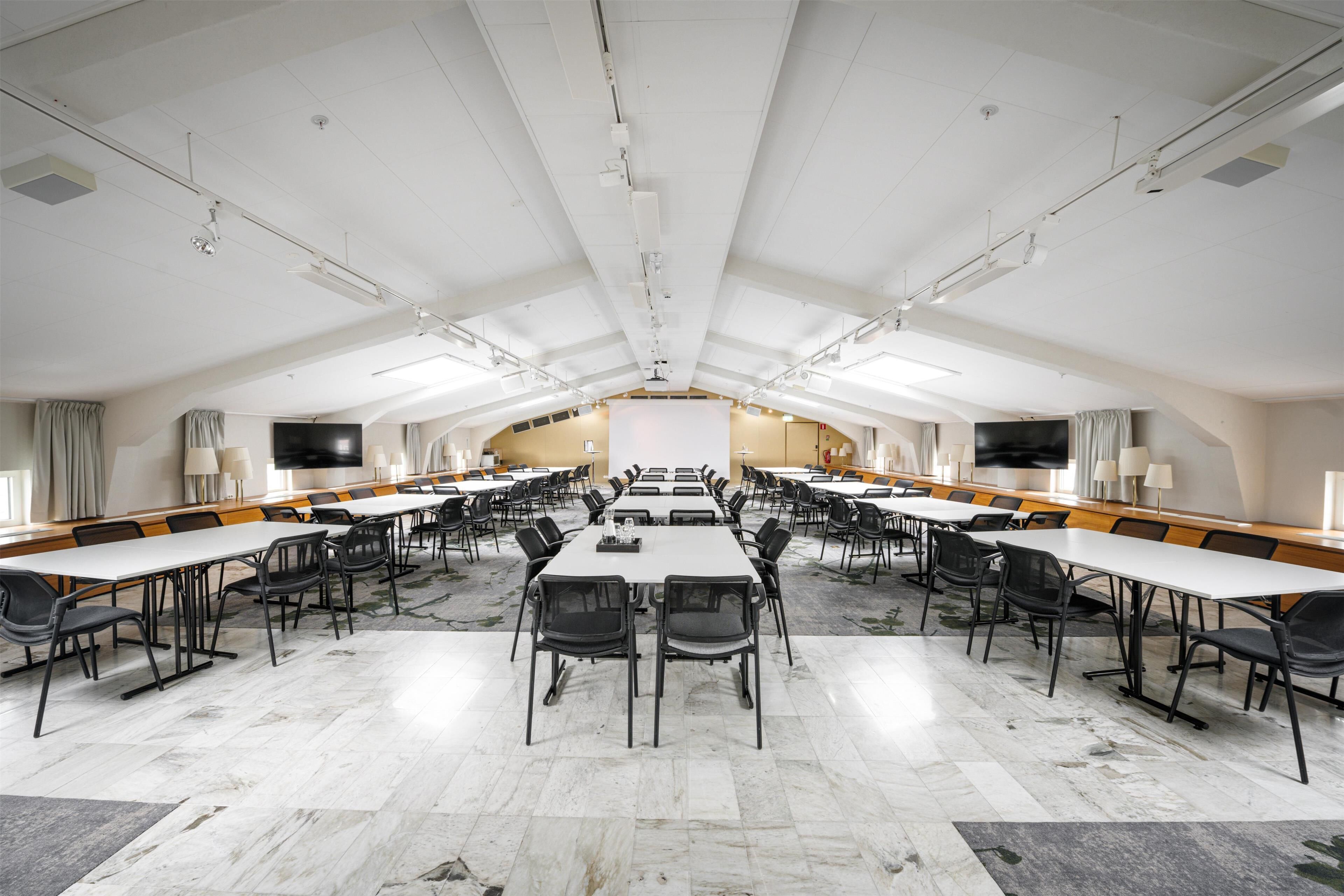 Un salón de conferencias amplio con mesas y sillas largas, una pantalla grande y un escenario al frente.