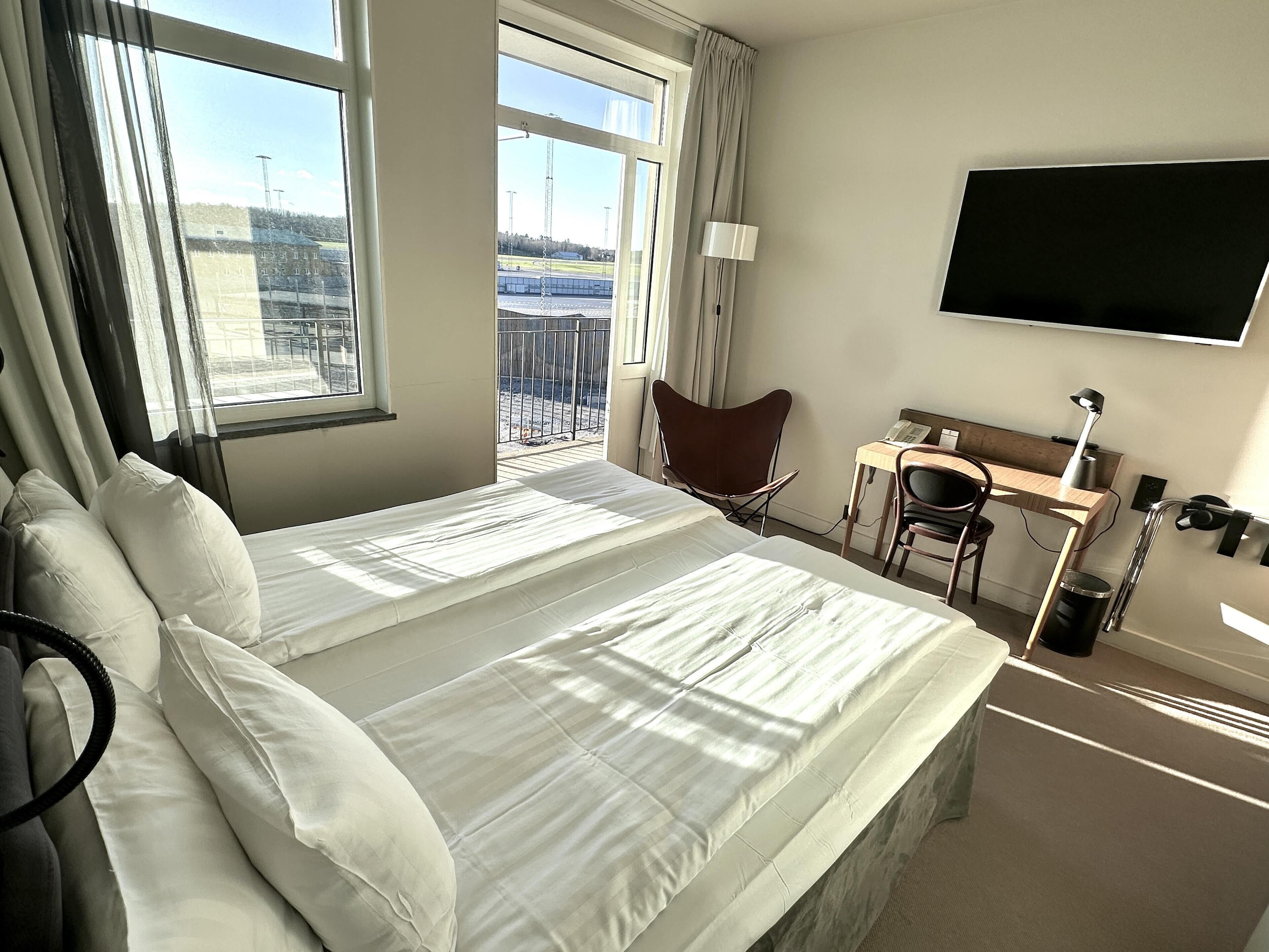 A hotel room with a large bed, a desk, a chair, a TV, and a view of the outside.