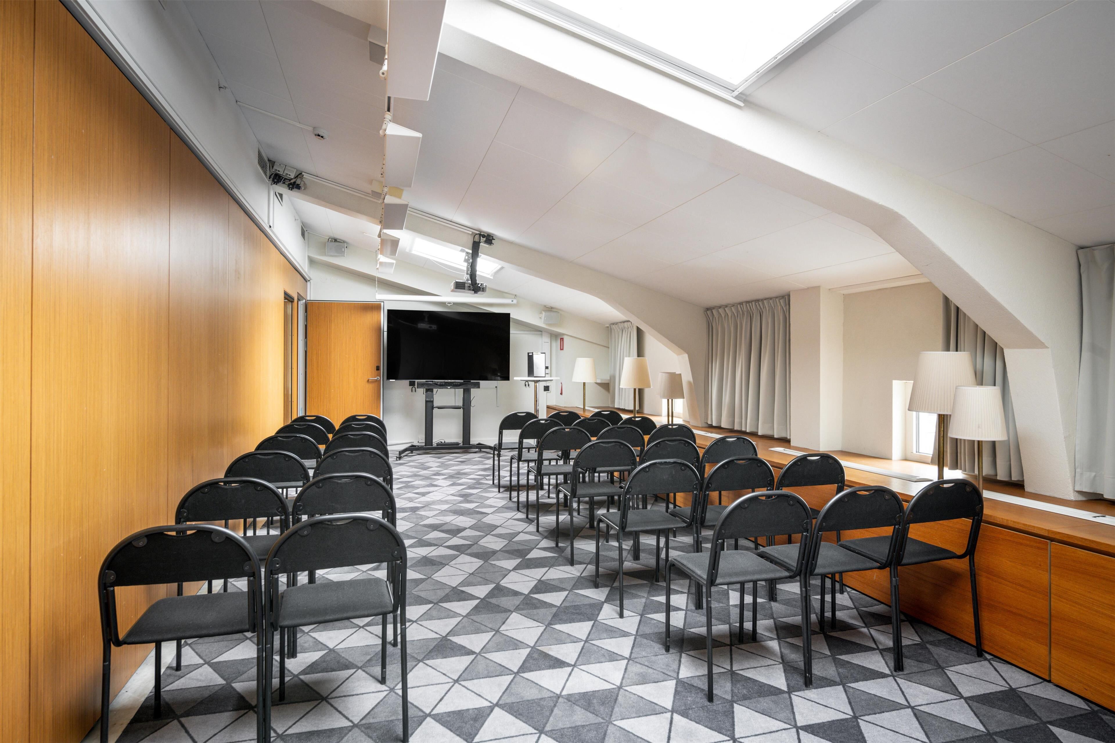Un salón de conferencias con suelo a cuadros, filas de sillas, una pantalla grande y una larga mesa con lámparas.