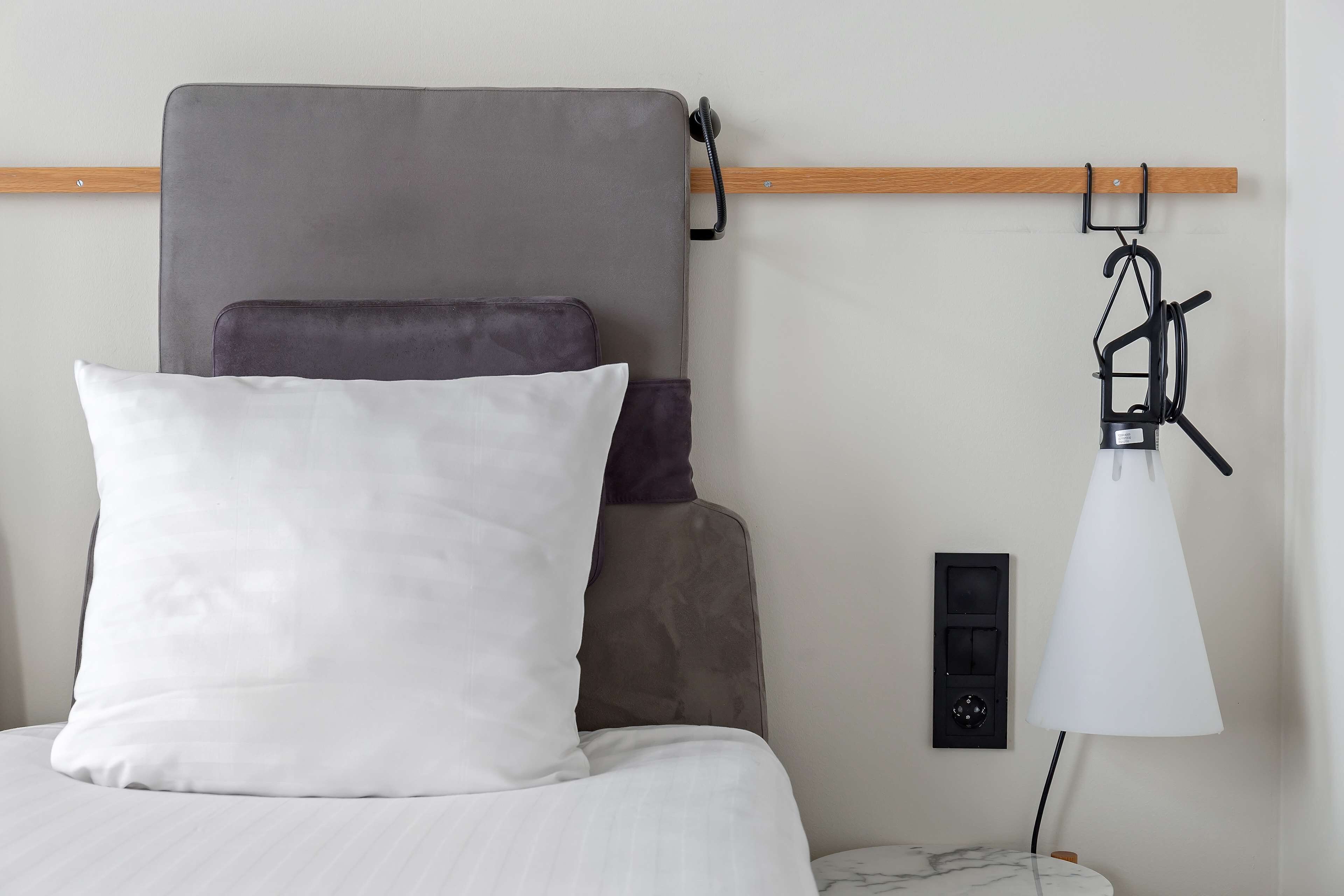 Una cama con cabecera gris, un almohadón blanco y un almohadón gris oscuro. Una lámpara blanca está sobre una mesita de noche.