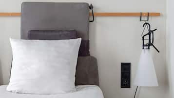 Una cama con cabecera gris, un almohadón blanco y un almohadón gris oscuro. Una lámpara blanca está sobre una mesita de noche.
