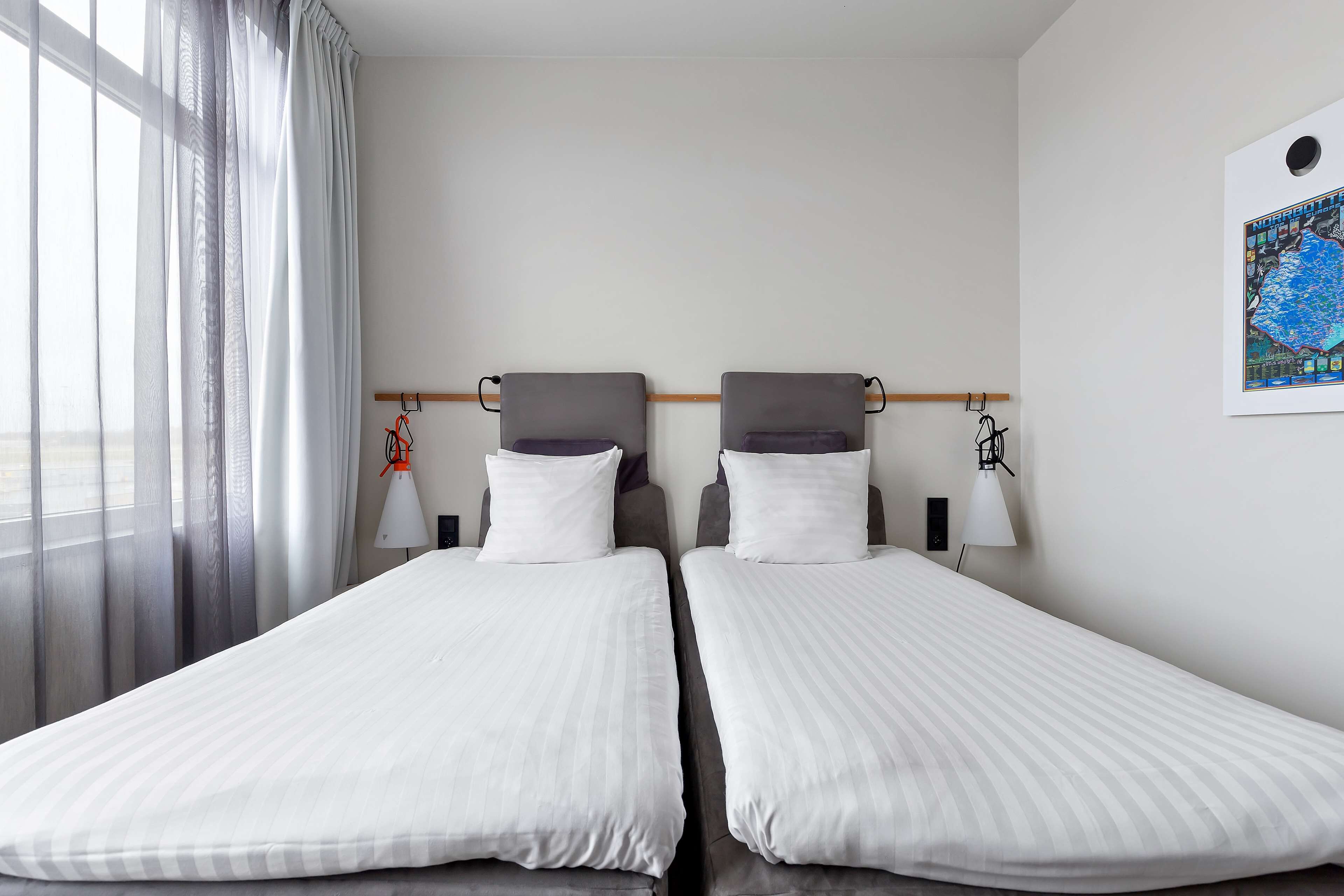 Habitación de hotel con dos camas individuales, una ventana con cortinas translúcidas, una lámpara de pared y un cuadro enmarcado.