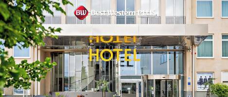 A hotel entrance with a glass facade, a sign for Best Western Plus, and a bicycle parked outside.