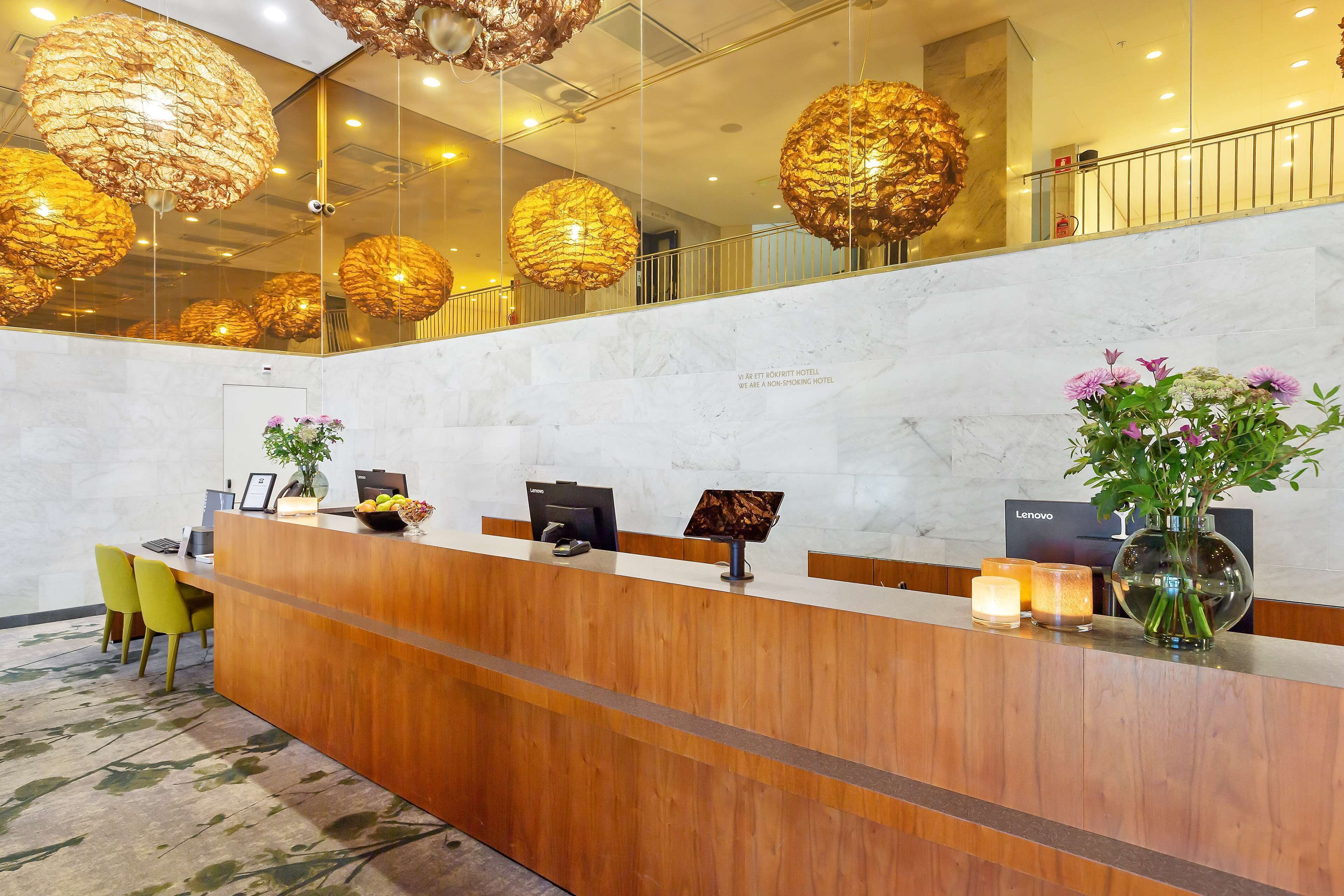 Un comptoir d’accueil d’hôtel avec un comptoir en bois, un ordinateur Lenovo, un vase avec des fleurs et un miroir qui reflète les luminaires du plafond et les éléments de décoration sphériques.