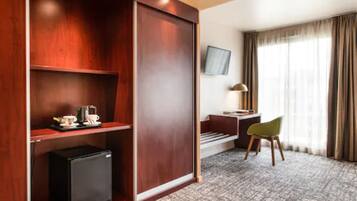 A hotel room with a wooden wardrobe, a desk with a chair, a television mounted on the wall, and a small refrigerator.