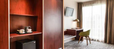 Habitación de hotel con un armario de madera, un escritorio con silla, televisor montado en la pared y un refrigerador pequeño.