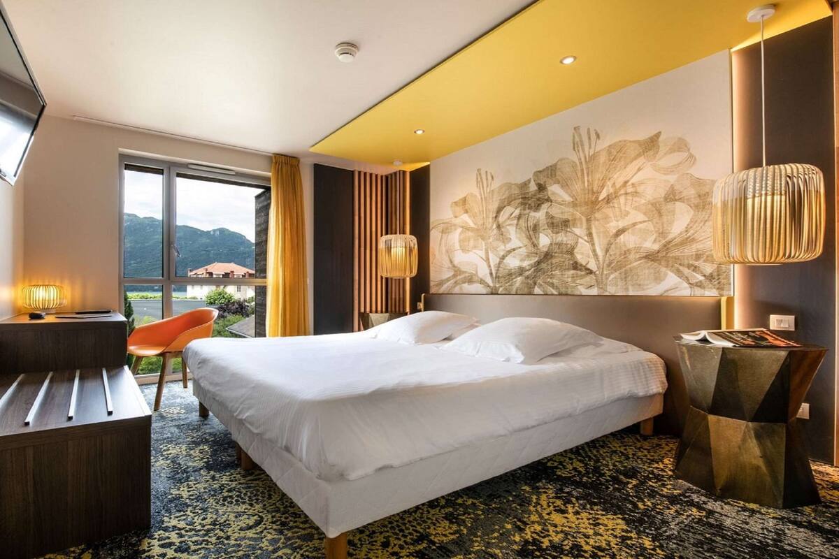 Une chambre d’hôtel avec un lit, un bureau, une chaise, une grande fenêtre et une vue sur les montagnes.