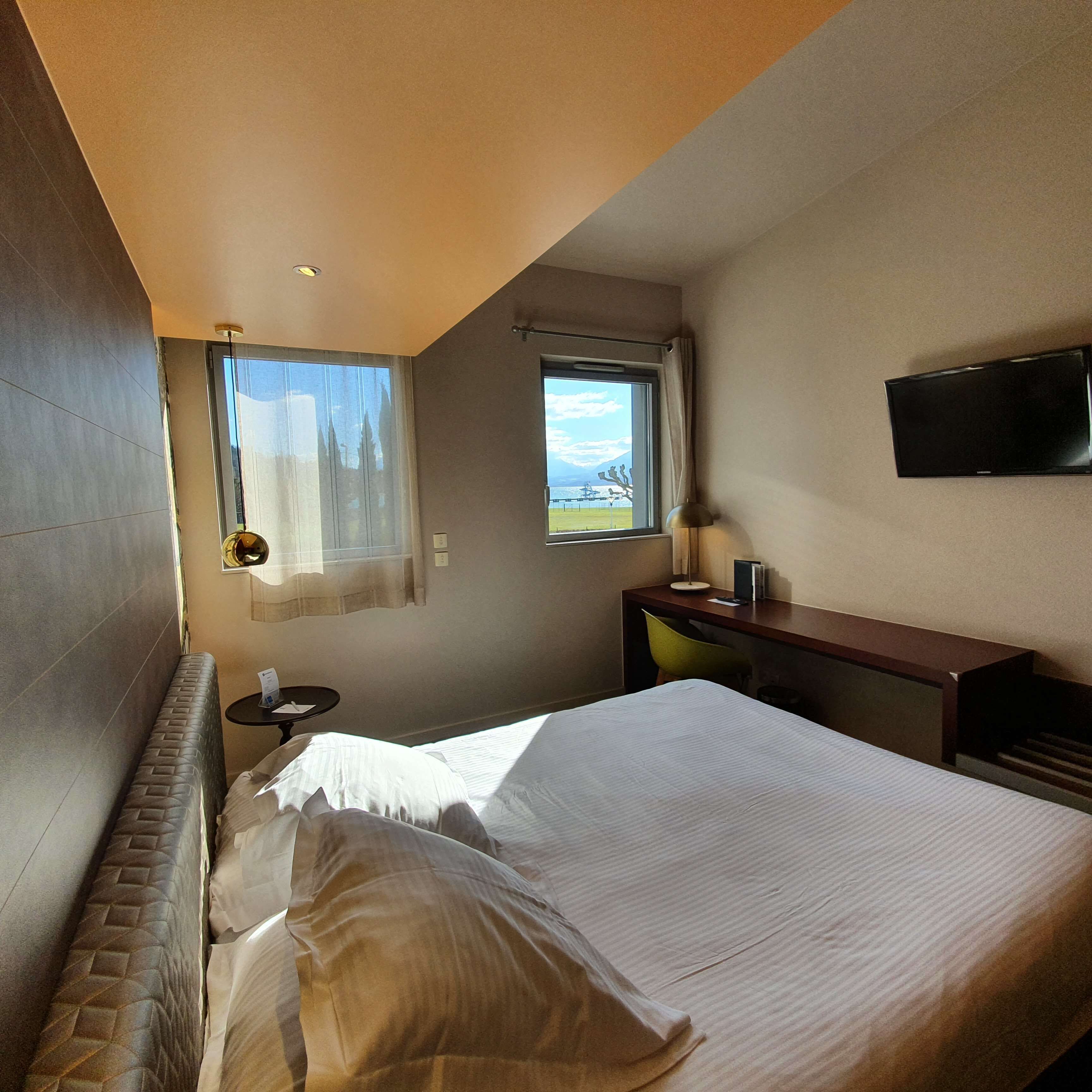 Hotellihuone, jossa on sänky, työpöytä, tuoli, televisio ja kaksi ikkunaa, joissa on verhot.