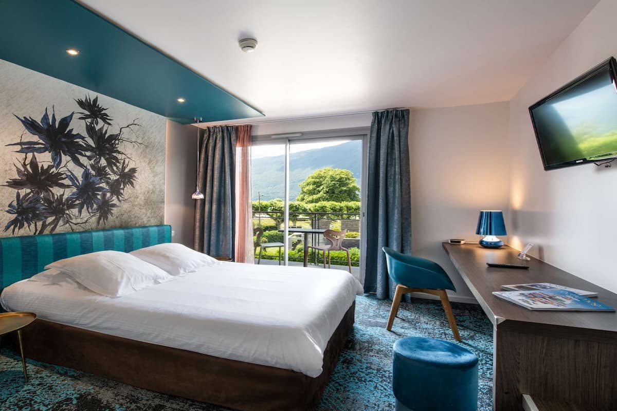 Une chambre d’hôtel équipée d’un lit, d’un bureau, d’une chaise, d’une télévision et d’une grande fenêtre donnant sur un paysage verdoyant.