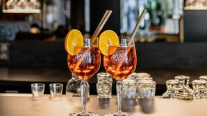 Deux verres contenant une boisson, chacun muni d’une paille et décoré d’une tranche d’orange, sont posés sur le comptoir d’un bar, avec des bouteilles et d’autres verres en arrière-plan.