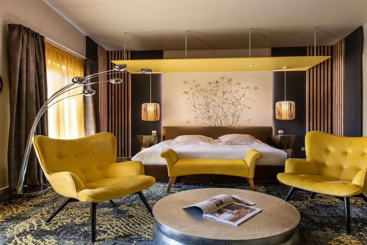 Une chambre d’hôtel avec un lit, deux fauteuils jaunes, une table basse ronde avec un magazine, une lampe sur pied et un mur décoré d’un motif représentant un arbre.