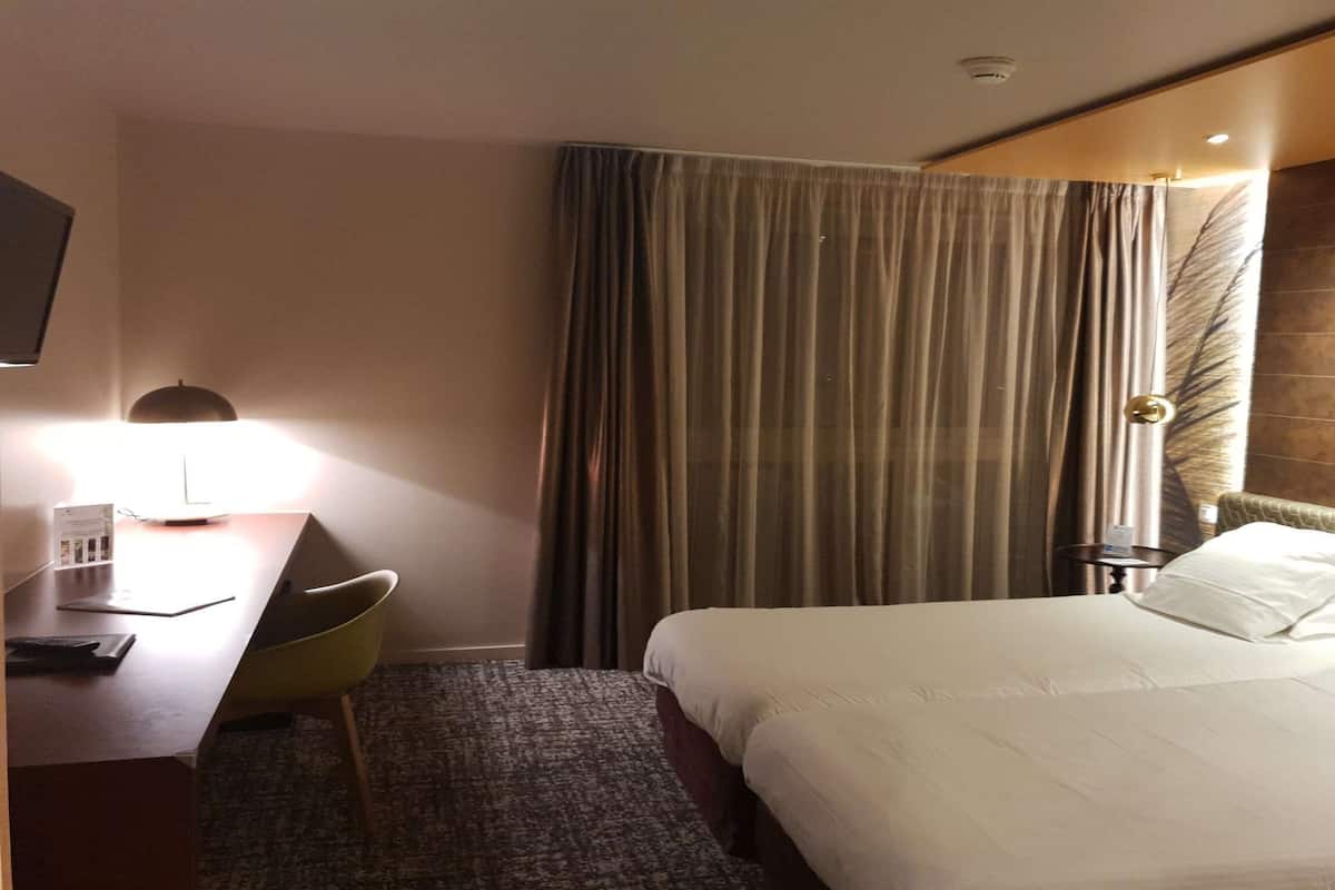Une chambre d’hôtel avec un lit, un bureau doté d’une lampe, une chaise et un rideau.