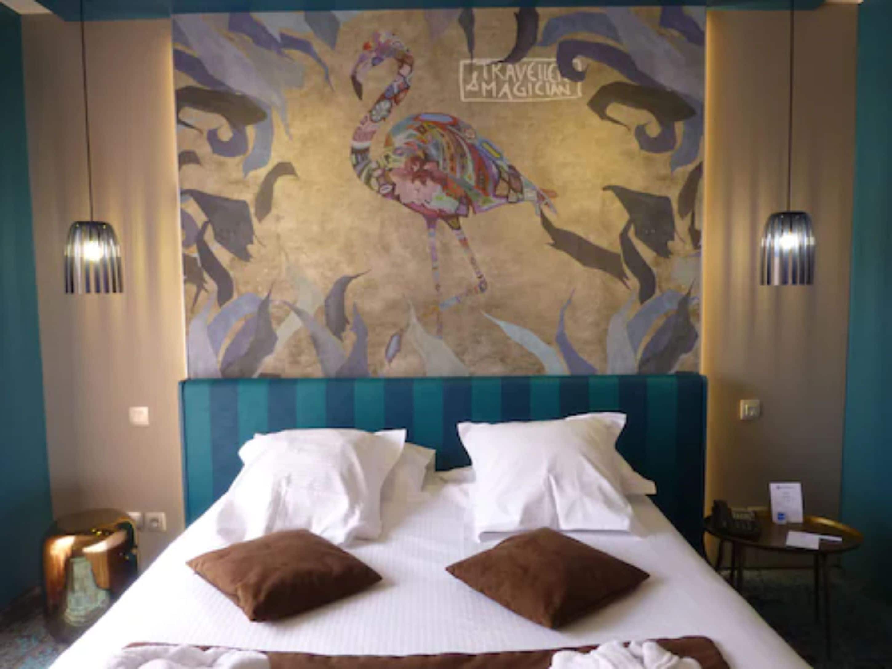A hotel room with a large mural of a colorful flamingo, a bed with white linens and two brown pillows, and two wall-mounted lamps.