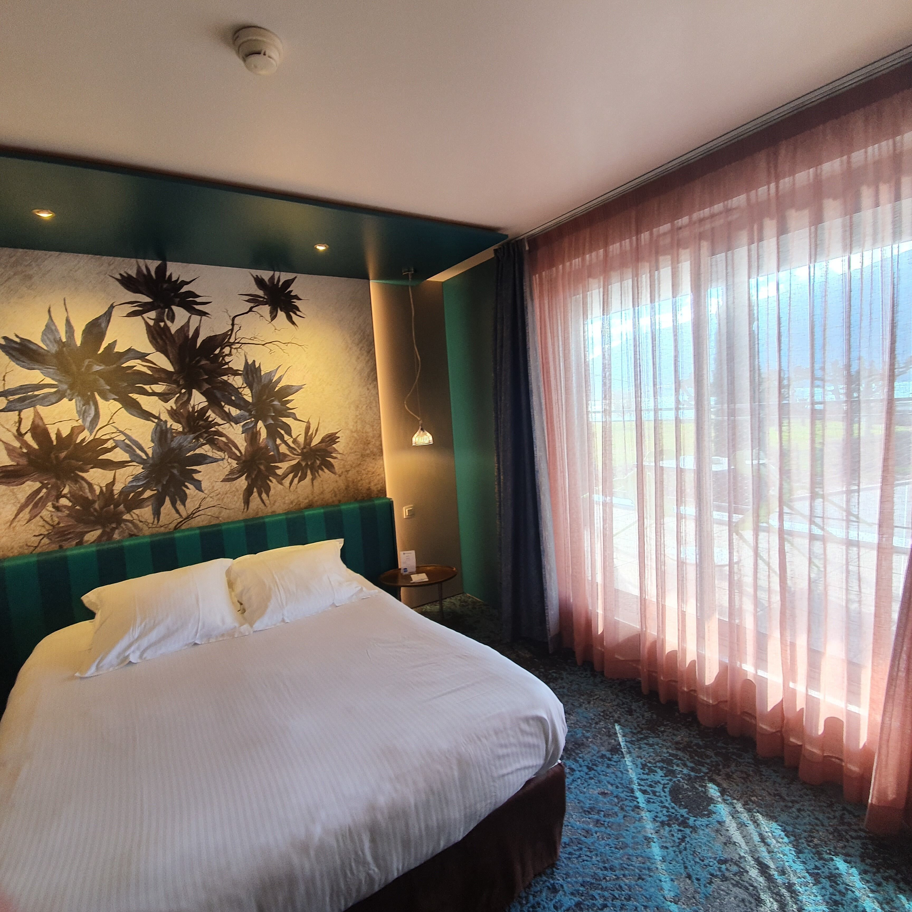 A hotel room with a bed, a wall mural, a window with sheer curtains, and a small table with a lamp.