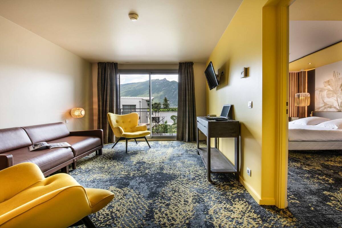 Une chambre d’hôtel avec un canapé marron, des fauteuils jaunes, un bureau avec un ordinateur portable, un lit et une vue sur les montagnes par la fenêtre.