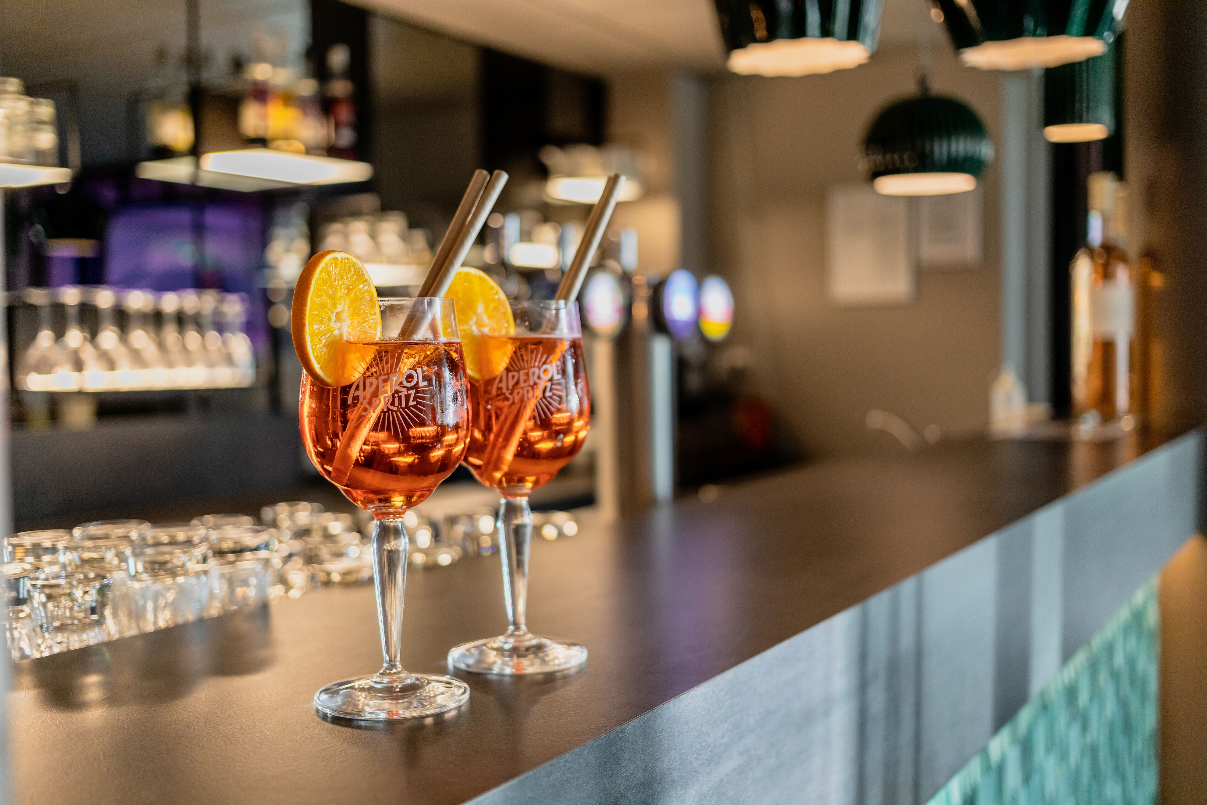 Two glasses of amber-colored beverage with orange slices and straws on a bar counter.