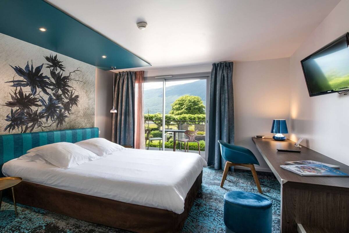 A hotel room with a bed, a desk, a chair, a TV, and a view of the outdoors.
