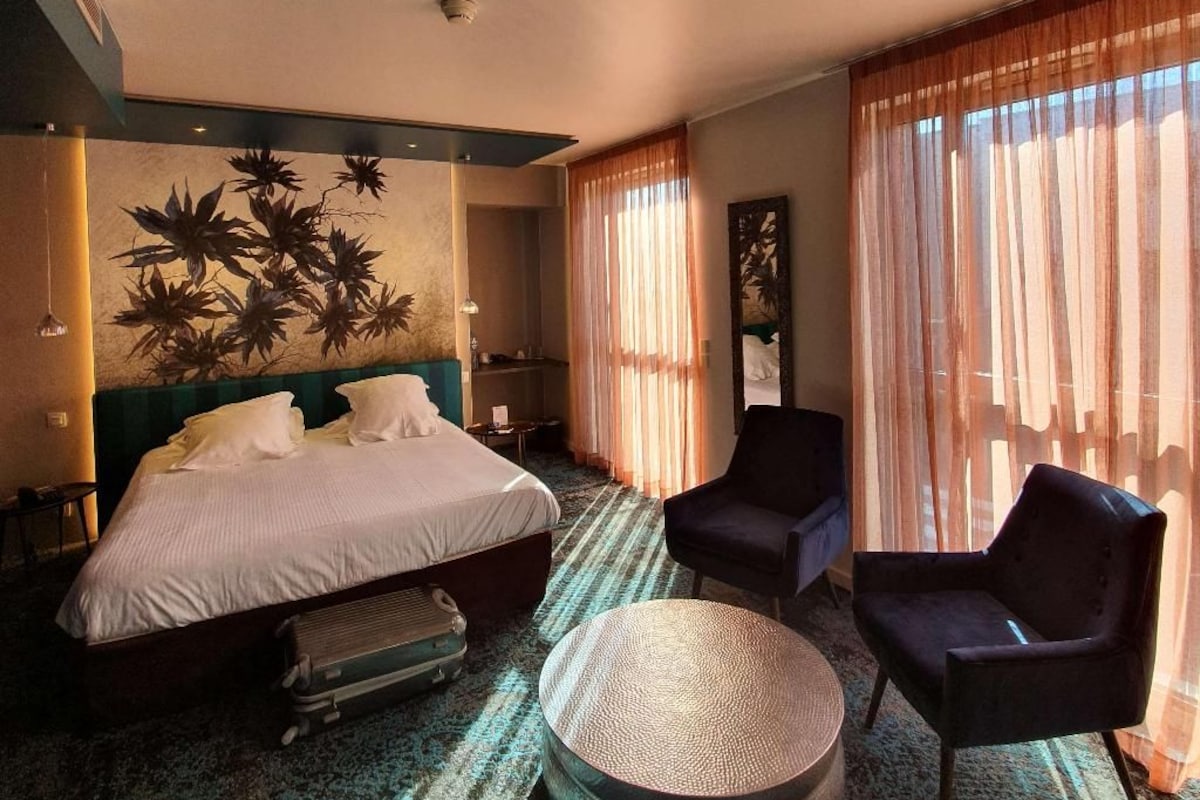 Een hotelkamer met een bed, een rond poefje, twee stoelen, een grote spiegel en een wand met een plantenmuur.