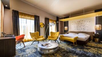 Una camera d'albergo moderna con un letto, due poltrone gialle, un tavolino rotondo, una scrivania con un portatile e un'ampia finestra con tende.