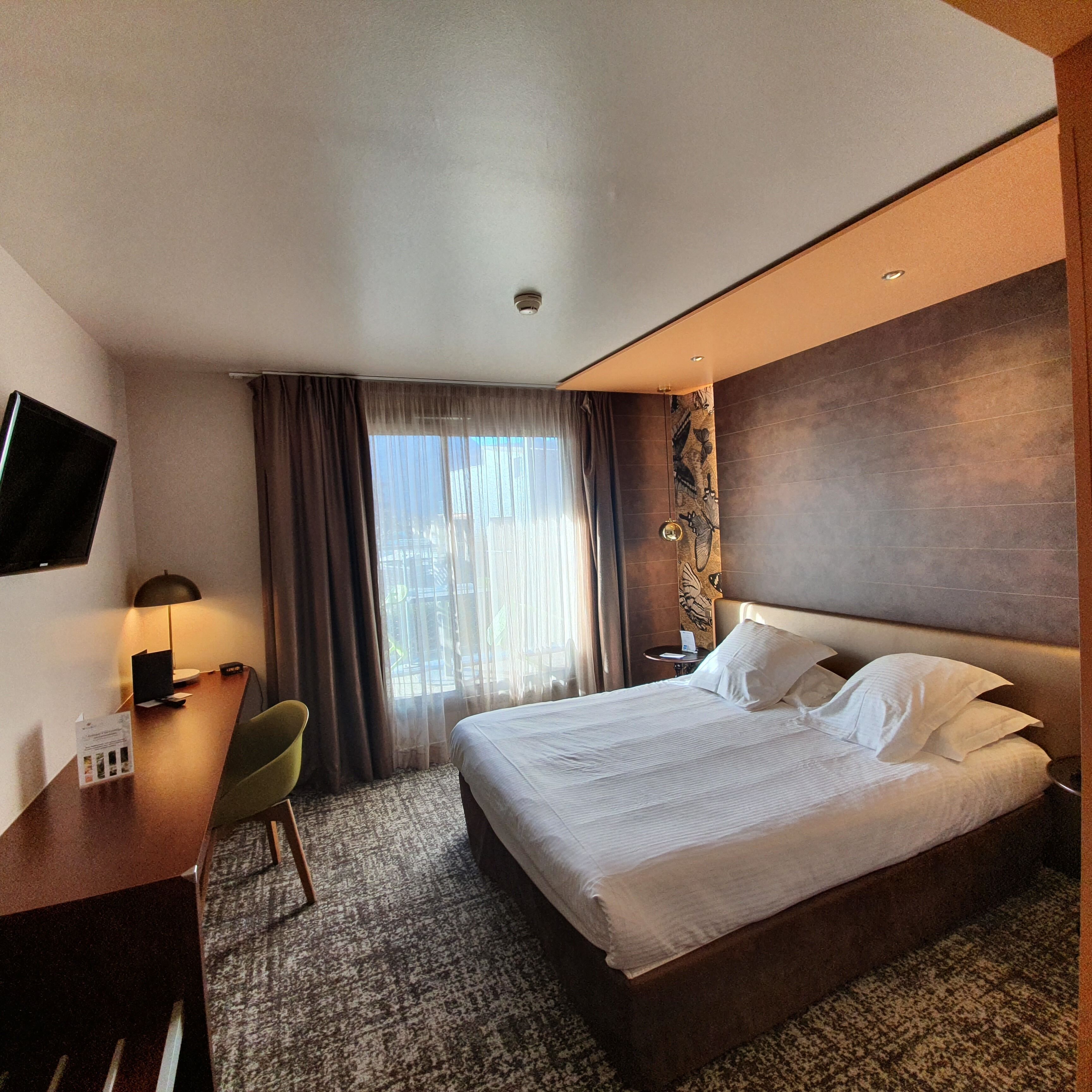 A hotel room with a bed, a desk, a chair, a TV, and a window with curtains.