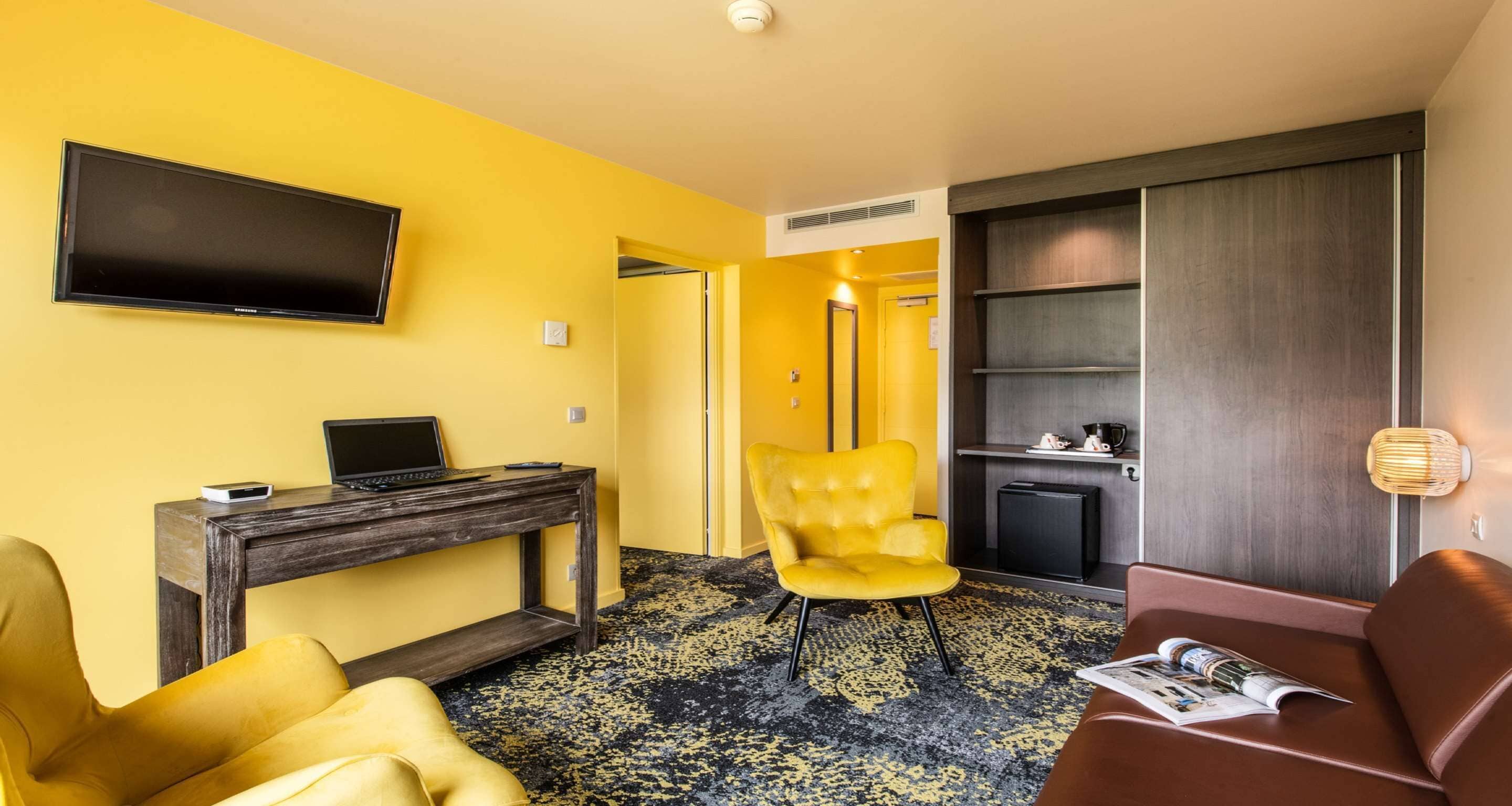 A hotel room with a yellow wall, a flat-screen TV mounted above a wooden desk, a yellow armchair, a brown sofa, and a magazine on the sofa.