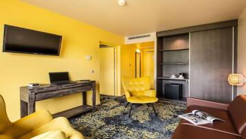 A hotel room with a yellow wall, a flat-screen TV mounted above a wooden desk, a yellow armchair, a brown sofa, and a magazine on the sofa.