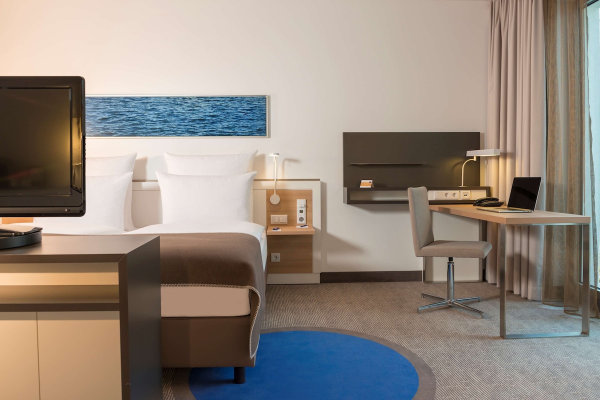 Een hotelkamer met een bed, bureau, stoel, televisie en een schilderij van de zee.