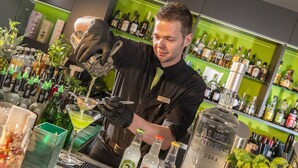 Een barman maakt een drankje klaar achter een bar, met flessen drank in de achtergrond.