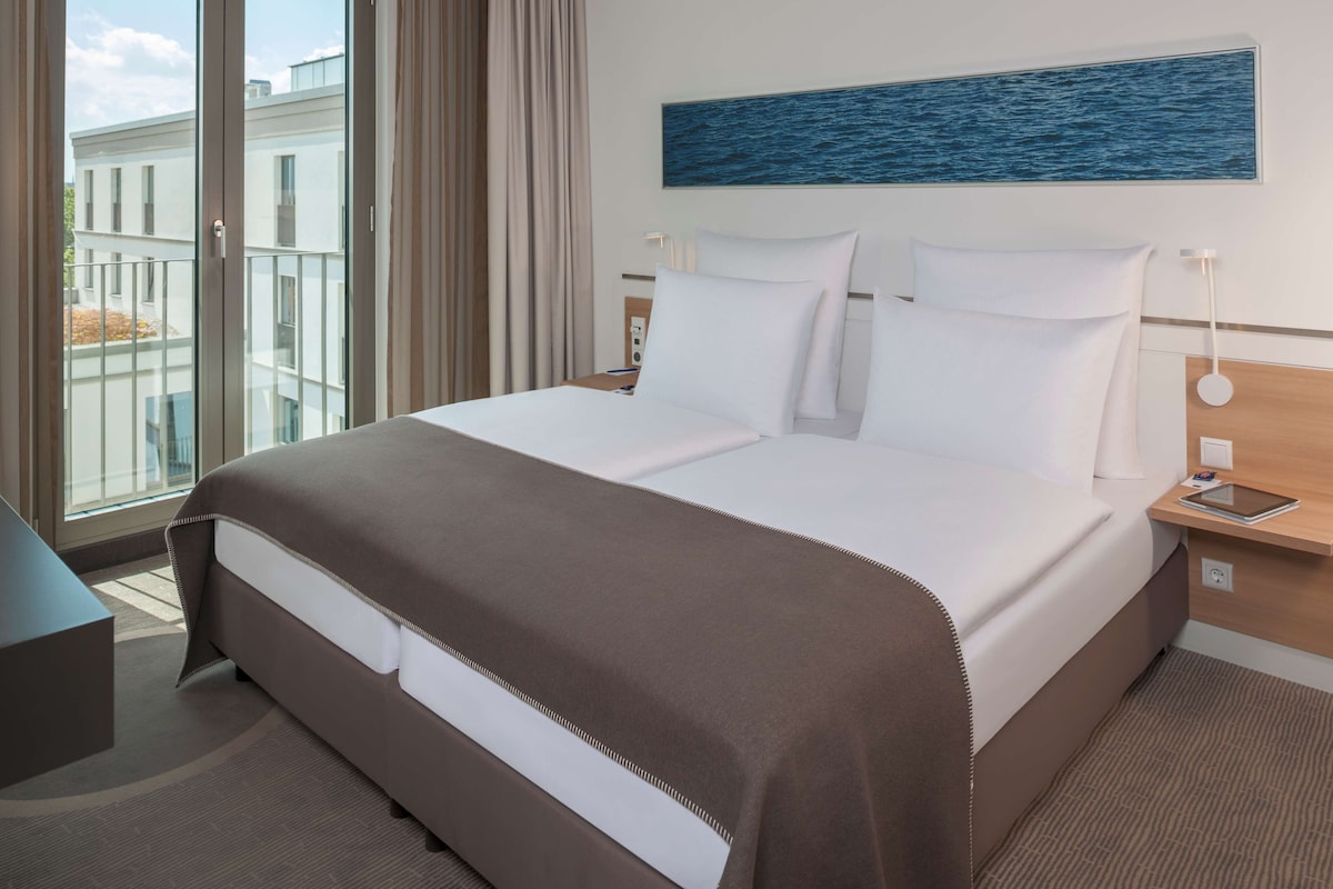 Hotelkamer met een groot bed, nachtkastje, raam met gordijnen en uitzicht op gebouwen.