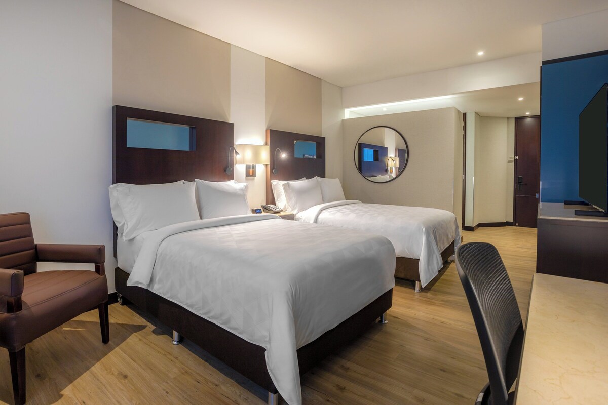 A hotel room with two beds, a desk, a chair, and a mirror.