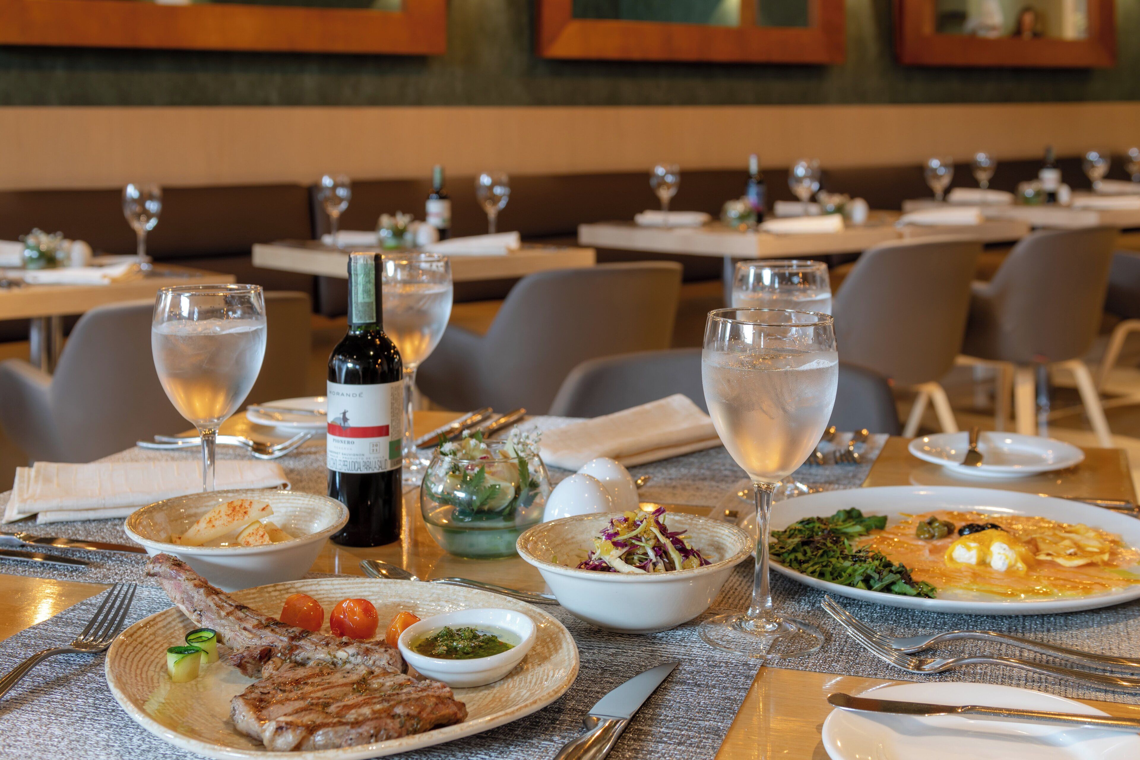 Ambiente de restaurante com mesas postas para refeição, pratos de comida, copos de água e uma garrafa de vinho.