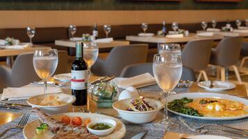 Ambiente de restaurante com mesas postas para refeição, pratos de comida, copos de água e uma garrafa de vinho.