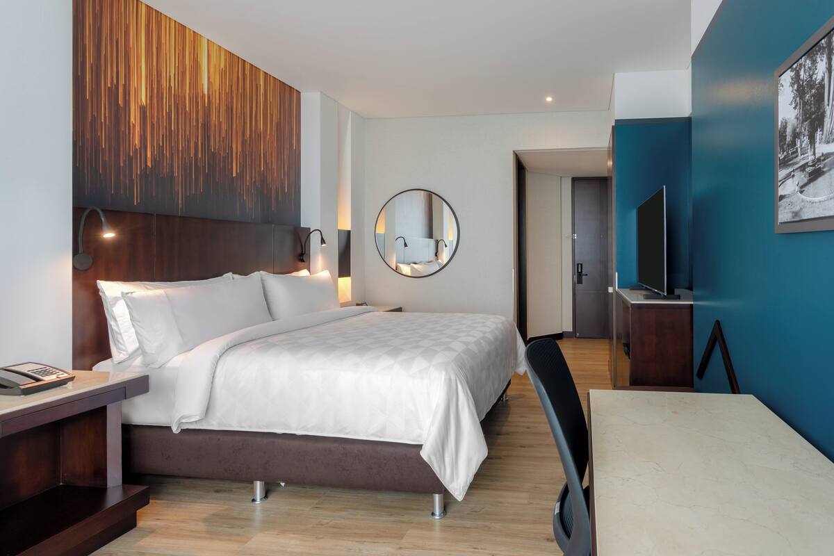 A modern hotel room with a large bed, bedside table, and a desk with a chair.