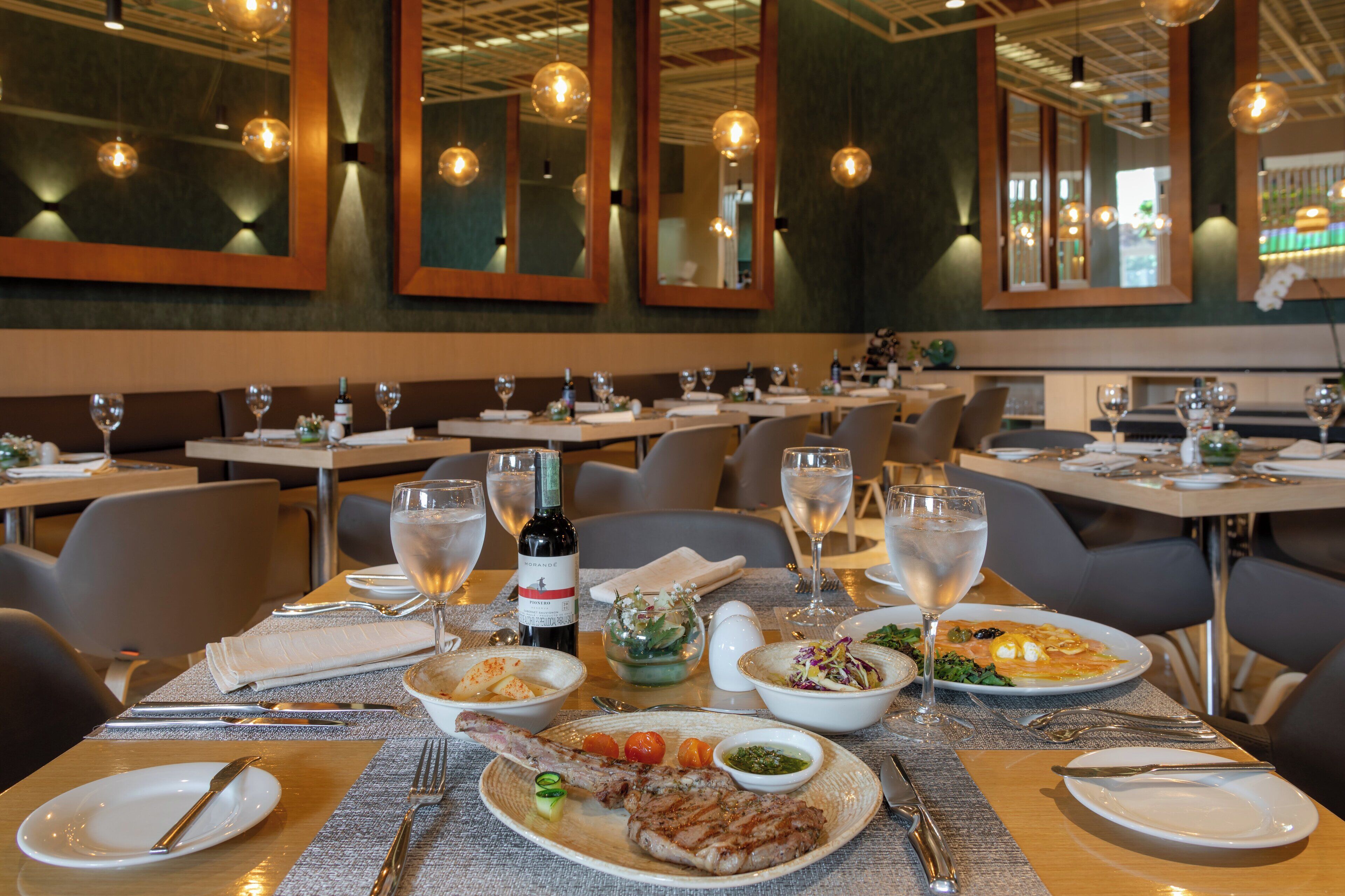 A restaurant interior with multiple dining tables set with plates, glasses, and bottles, and a variety of dishes including a steak and salad.