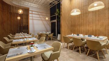 A modern dining area with wooden panel walls, multiple tables with chairs, and hanging pendant lights.