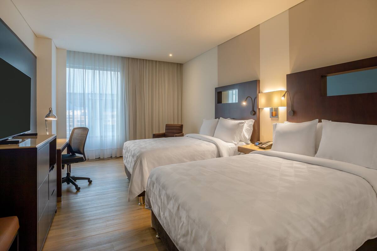 A hotel room with two beds, a desk, a chair, a TV, and a large window with curtains.
