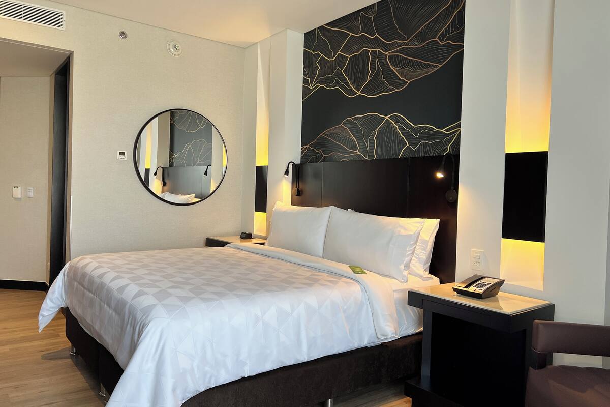 A modern hotel room with a large bed, a round mirror, a headboard with a leaf pattern, bedside lamps, and a phone on the nightstand.