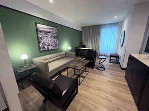 A modern living room with a sofa, a coffee table, a black chair, a small round table, and a desk with a chair. There is a large framed picture on the wall, a lamp on a side table, and a window with curtains.