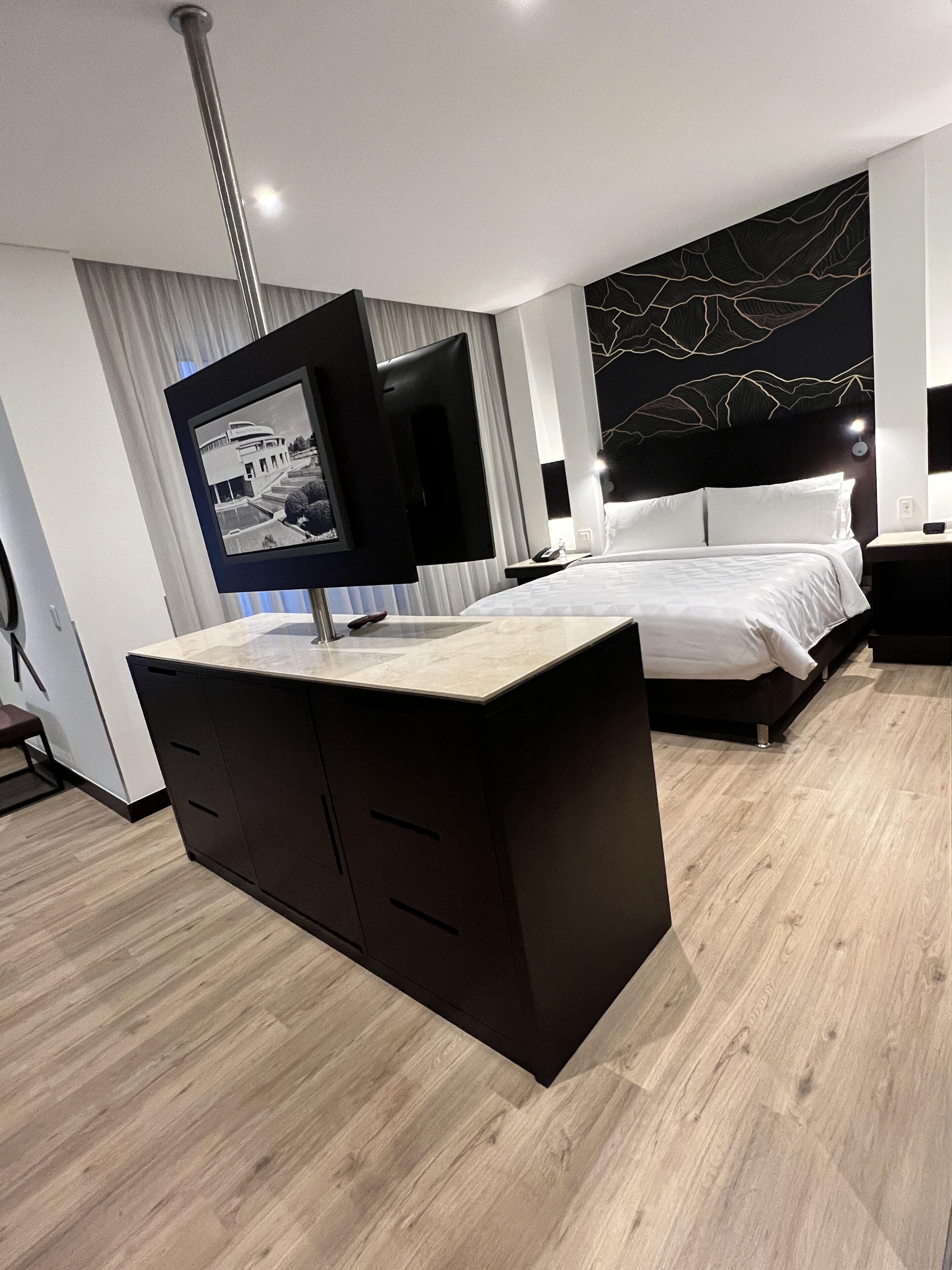 A modern hotel room with a large bed, a bedside table, a desk with drawers, a flat-screen TV mounted on the wall, and a decorative wall piece.